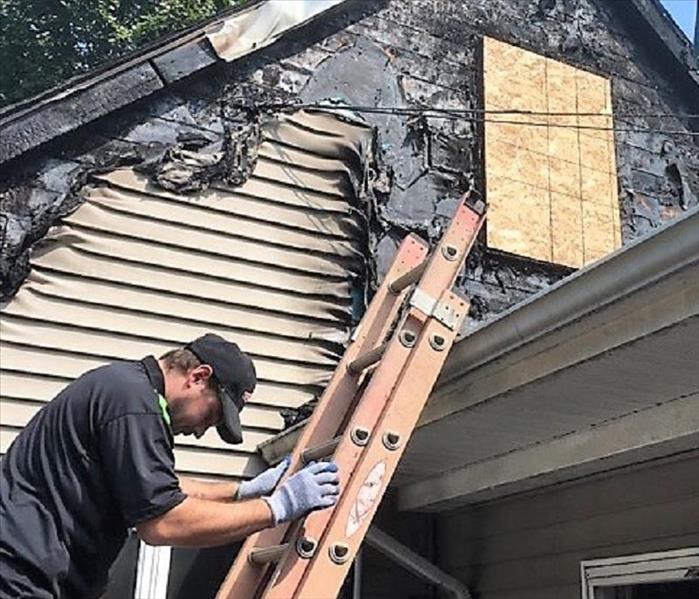Man climbing down a ladder of a house with melted siding and wood over the window